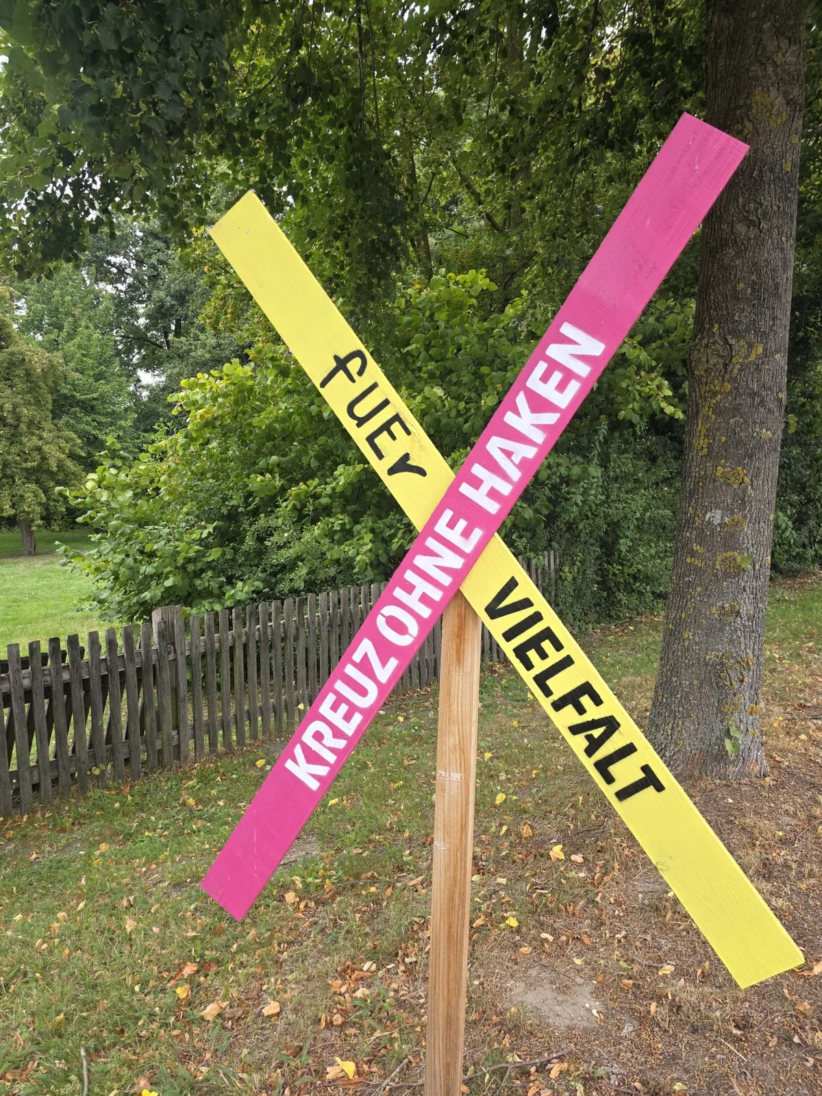 Kreuz ohne Haken fUEr Vielfalt, pinkes und gelbes Holzkreuz am Gartenzaun
